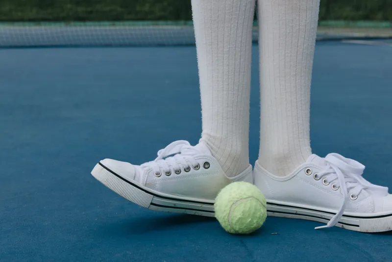 A close up of a child s feet wearing white sneakers and knee high socks, standing on a blue tennis court next to a green tennis ball, embodying enthusiasm and playfulness