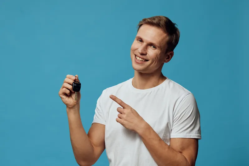 Smiling man holding car key pointing at it, casual white t-shirt, cheerful expression, blue background, studio portrait, lifestyle concept, positive emotion, holding vehicle key, happy man.