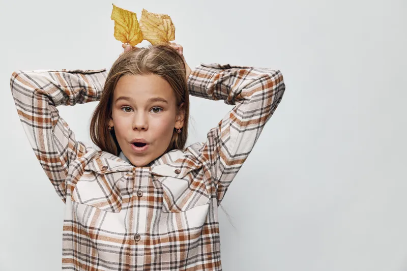 Girl in plaid shirt holding fallen autumn leaves behind head on white background with surprised facial expression, seasonal mood and playful gesture for fall concept.
