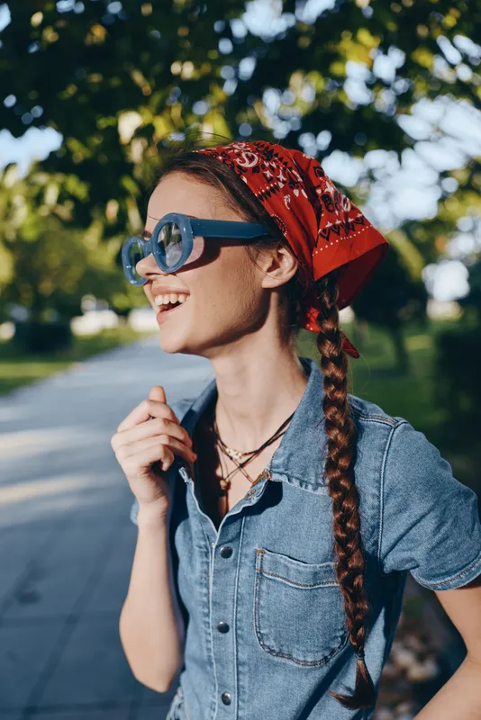 Lifestyle woman wearing modern boho-western denim street style with a red bandana and sunglasses, smiling outdoors in natural light, film color aesthetic.