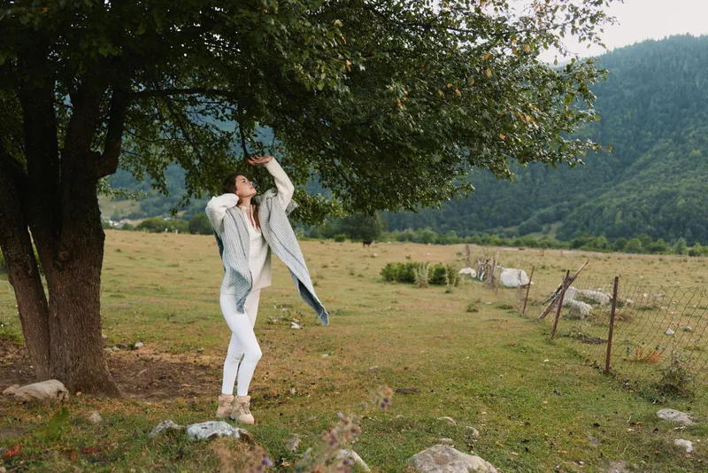 Young woman in a light coat and white jeans leaps playfully under a large tree in a rural field, capturing carefree movement and outdoorsy joy against rolling hills and grazing sheep, conveying