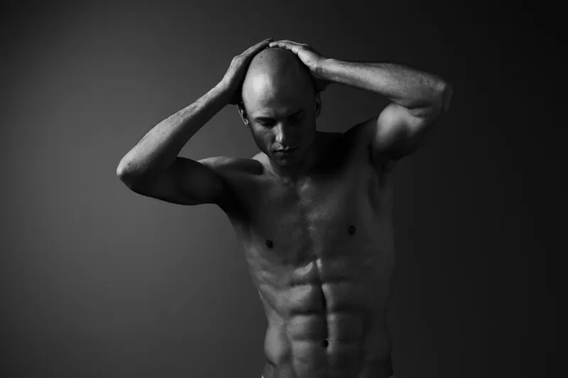 Black and white image depicting a focused man, showcasing his strong physique while his hands grasp his head, reflecting deep emotion and strength.