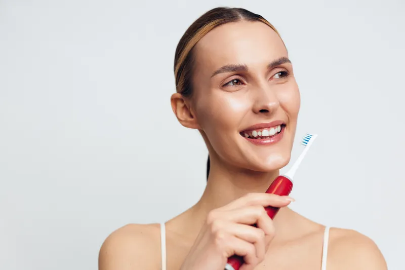 Young woman with a bright smile holds a red toothbrush near her face, wearing a white tank top, isolated against a soft light background.