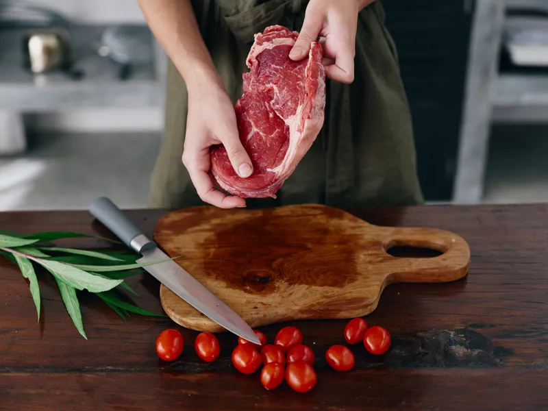 Женщина с ножом в руке нарезает свежее стейковое мясо для жарки на кухне, на столе соль, перец и другие специи, красные помидоры черри и зелень, приготовление ужина