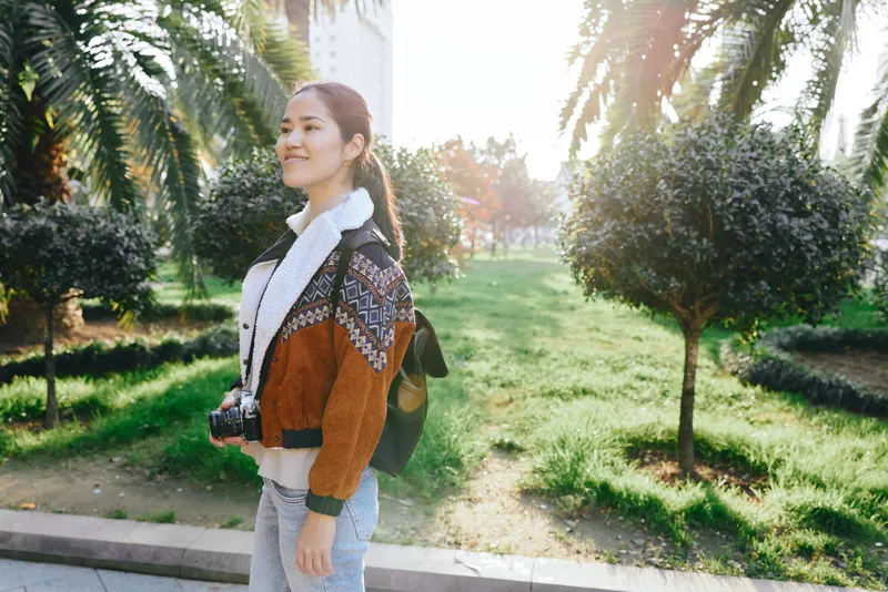 Woman with camera in a sunny park, wearing cardigan and scarf, strolling along a green path, trees and light flare creating a relaxed outdoor lifestyle scene for stock