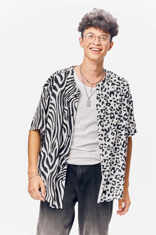 Young man wearing a stylish black and white patterned shirt with glasses smiling confidently, isolated on a light background showing casual street fashion and modern trends.