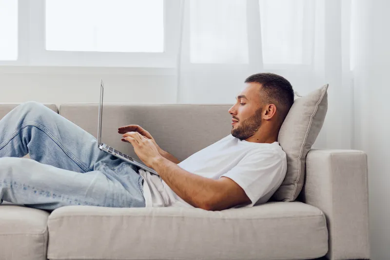 Relaxed man working on a laptop at home, wearing casual clothes, the cozy setting enhances productivity and comfort. Ideal for modern lifestyle themes.