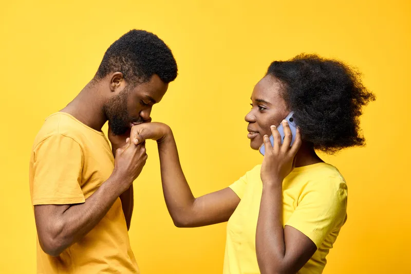 Young African couple sharing a joyful moment, with the man kissing the woman s hand while she talks on the phone in bright yellow attire against a vibrant yellow background