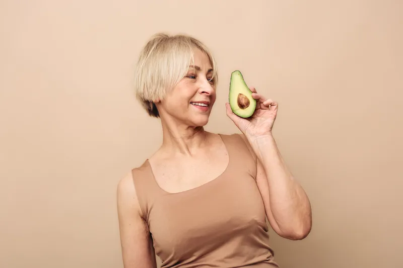 Smiling mature woman with short blonde hair holds fresh avocado half near face against beige background. Concept of healthy eating, nutrition, wellness, and natural lifestyle for seniors.