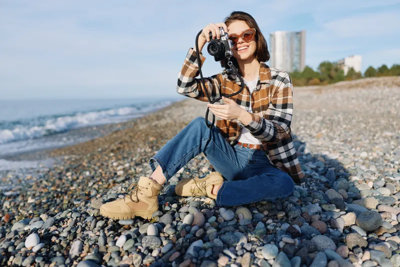 Photographer woman on rocky beach posing with camera, capturing seaside landscape and sunlight, outdoor lifestyle shot with waves, stones and blue sky