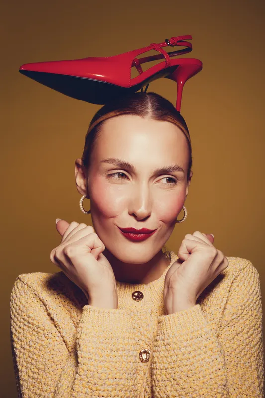 Woman with red high heel shoe balanced on her head wears yellow textured sweater and gold hoop earrings. Studio portrait with warm lighting and playful expression on brown background.