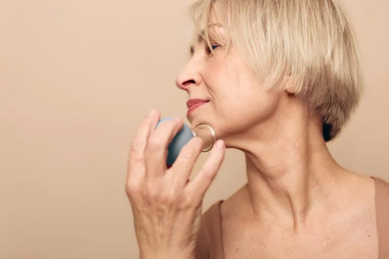 Mature woman with short blonde hair applying perfume on neck, close-up profile on beige background. Concept of personal care, elegance, fragrance, beauty routine, and self-confidence.