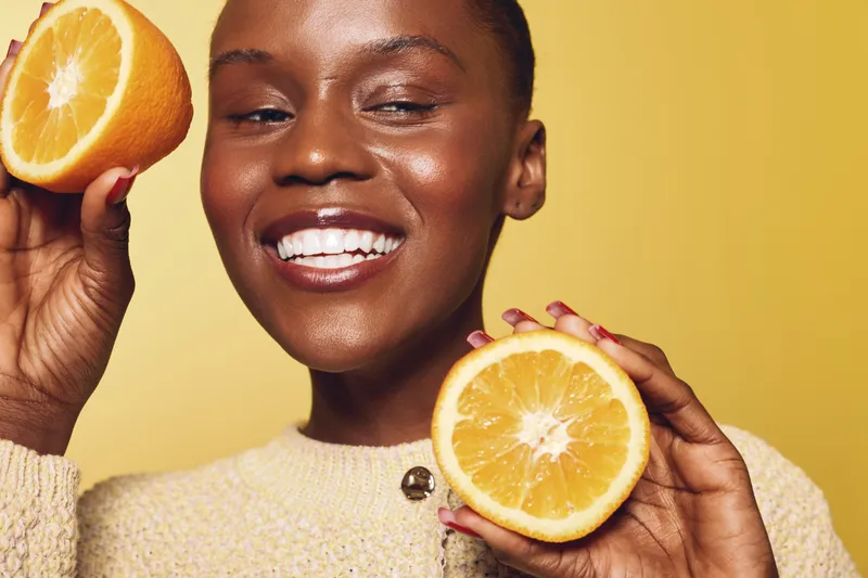 Smiling young black woman with dark skin holding two halves of fresh orange fruit. Close-up on yellow background. Concept of healthy lifestyle, natural beauty, and vitamin c benefits.