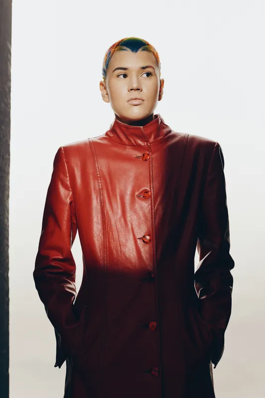 Woman with buzz cut wearing a modern red leather coat, posing confidently against a white background with strong dramatic lighting and shadow contrast.