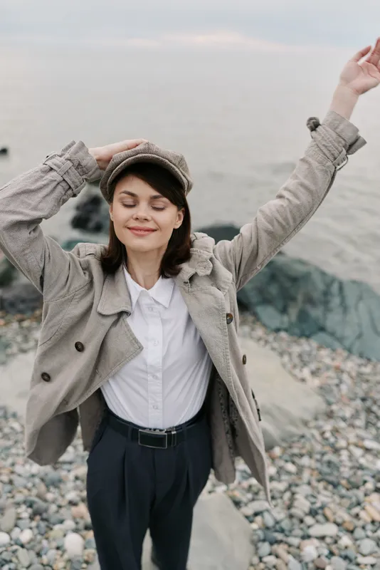 Smiling woman in stylish jacket and hat stands on pebble beach with eyes closed and arms raised. Concept for happiness, freedom, nature, and relaxation by the sea.
