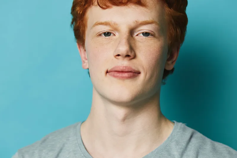 Portrait of young man with red hair and freckles wearing gray shirt on blue background. Closeup face, natural expression with casual style and soft lighting.