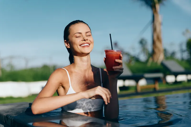 Poolside woman in a swimsuit drinks a refreshing beverage, basking in sunshine and outdoors, conveying vacation mood and summertime relaxation with happiness