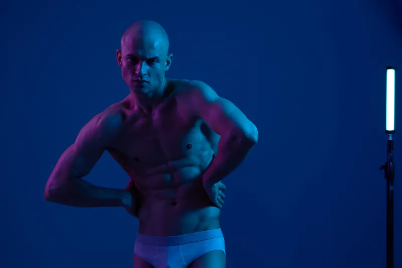 Athletic man posing confidently under vibrant blue lighting, showcasing his toned physique in an artistic black and white image.