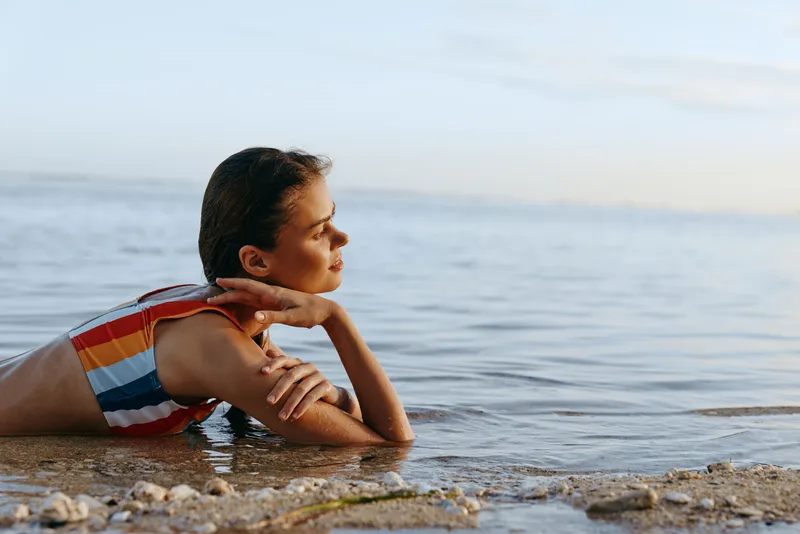 A young woman with brunette hair in a colorful striped swimsuit relaxes lying on her stomach at the edge of a calm sea on a pebble beach during a peaceful moment.