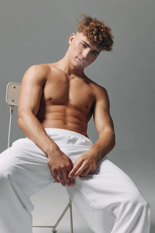 Young man with curly hair and muscular toned body sitting casually on chair wearing white pants against plain gray background in studio lighting for fitness portrait concept.
