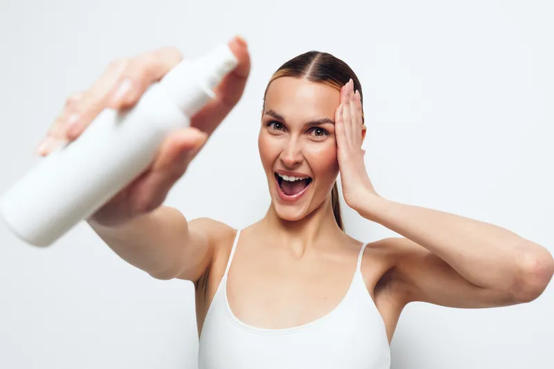 Smiling young woman sprays facial mist on her face with one hand touching her cheek isolated on white background skincare routine beauty concept.