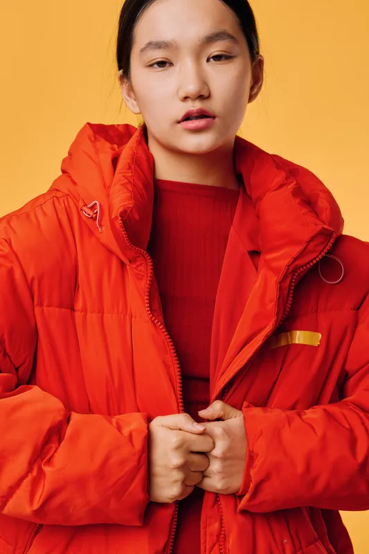 Young woman in orange puffer jacket standing against yellow background looking at camera with calm expression. Female model holding jacket edges wearing casual warm clothing for cold weather.