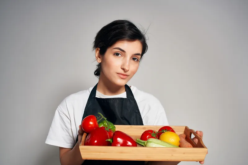 Домохозяйка в черном фартуке на фоне свежих овощей и продуктов