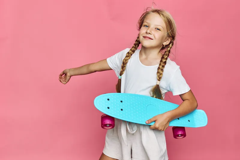 Smiling young girl with braids holding a blue skateboard on a pink background, wearing a white t-shirt and beige shorts.