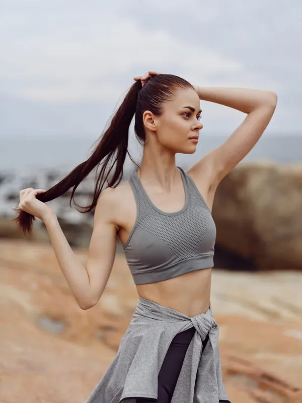 A young athletic woman with long dark hair pulls her ponytail back, wearing a grey sports bra and leggings, against a scenic coastal backdrop, showcasing fitness and confidence