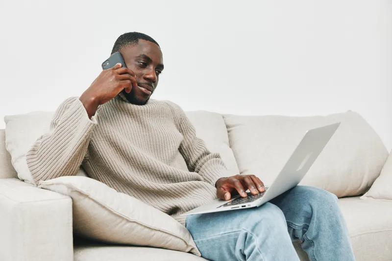 Young black man in cozy sweater working on laptop and talking on smartphone at home, conveying positivity and productivity in a modern setting