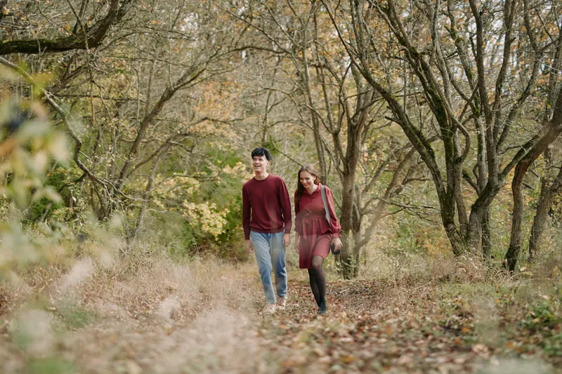 A young couple strolls along a leaf-strewn forest path in autumn, sharing a candid moment that conveys authenticity and realism through natural poses, soft light and muted tones.