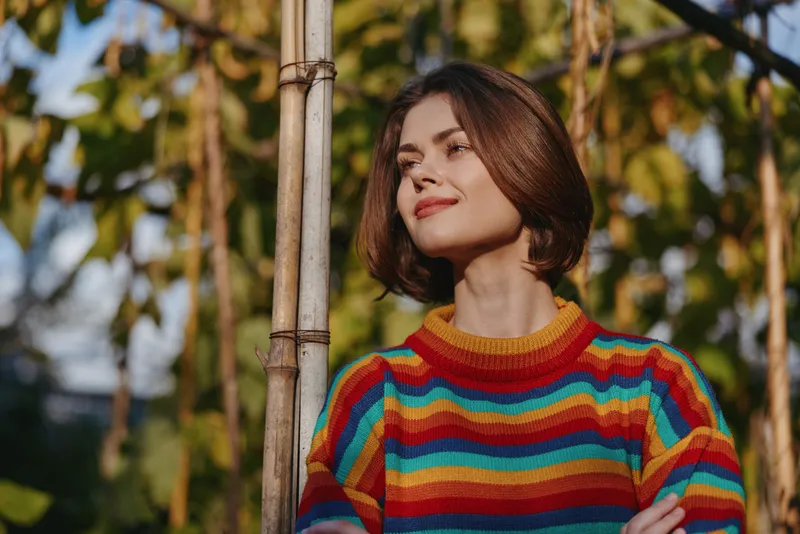 Woman in a colorful striped sweater with short hair leaning against bamboo in a tropical outdoor portrait, smiling gently in a casual outfit for lifestyle and travel fashion imagery.