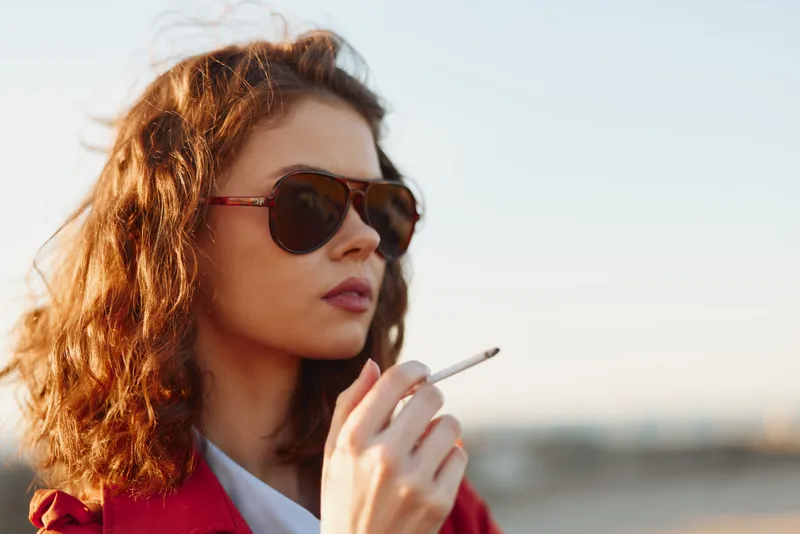 Woman with wavy hair wearing sunglasses, smoking a cigarette outdoors. Warm light and city backdrop convey fashion, mood, and casual confidence.
