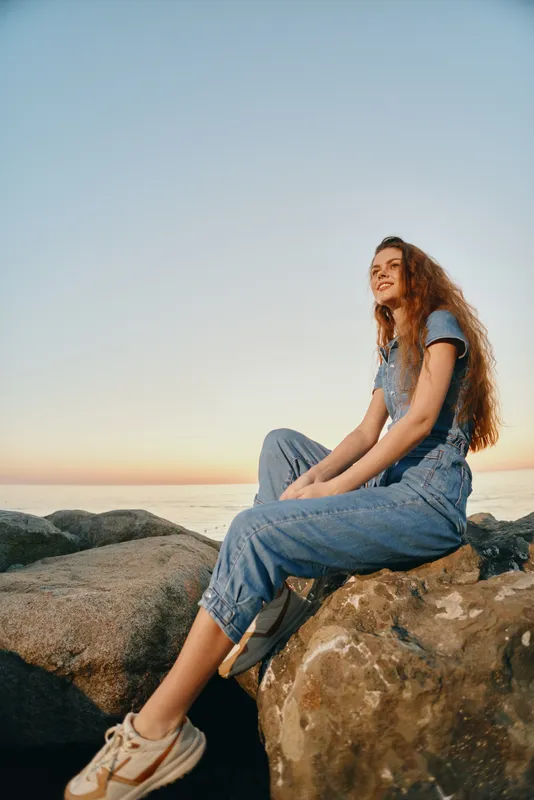 lifestyle woman in modern boho-western denim street style sitting on rocks at sunset, relaxed mood, film color tones, casual outdoor fashion