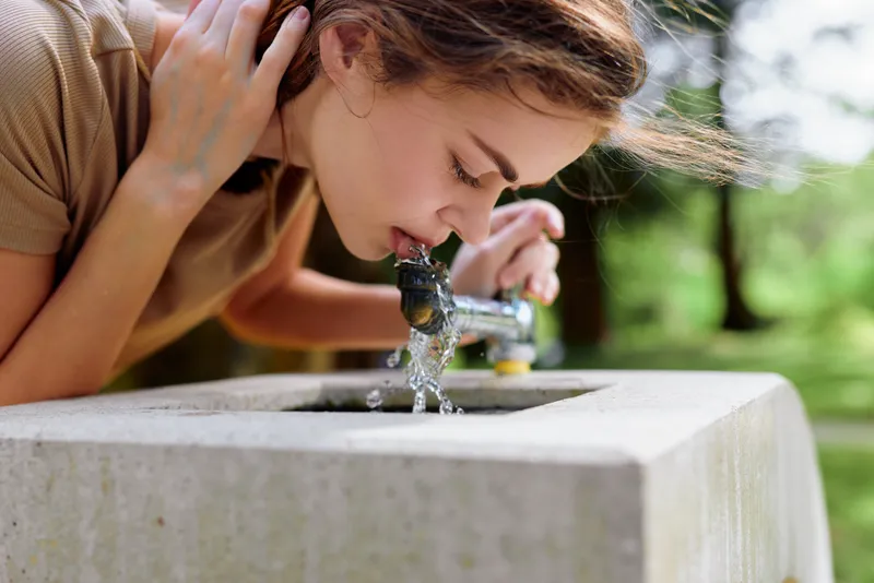 Woman drinking water from an outdoor fountain tap in a park, portrait closeup of young female leaning to drink, natural sunlight and splashing motion conveying refreshment.
