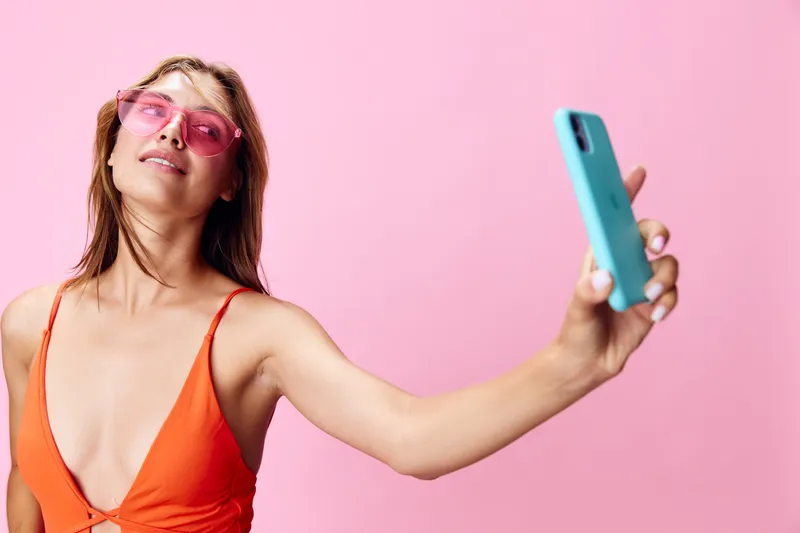 Модная молодая женщина в купальнике делает селфи на смартфон на ярком розовом фоне
