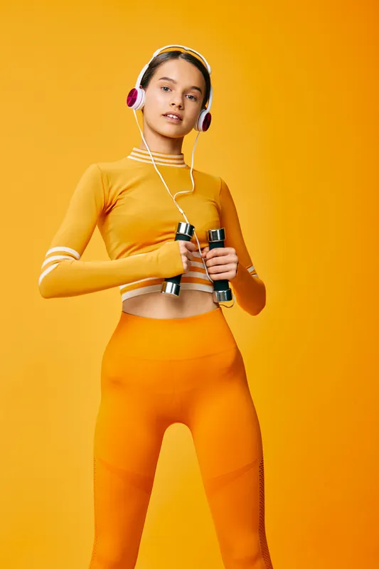 Young woman fitness portrait in orange activewear holding dumbbells and wearing headphones against a seamless orange studio background, confident sporty pose and healthy energy.