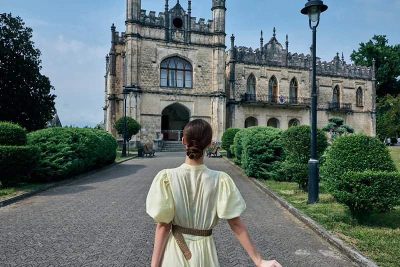 Woman in vintage dress walks toward a historic castle, lush gardens lining a gravel path, creating a timeless travel moment, as sunlight glints on stone and hedges frame the scene with quiet elegance