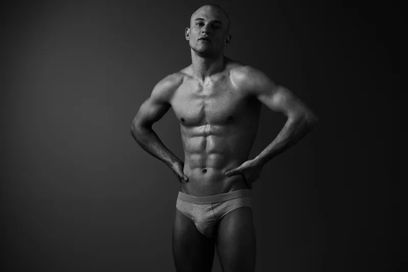 Athletic man posing confidently in black and white, showcasing strong physique with dramatic lighting, creating an impactful visual presence.