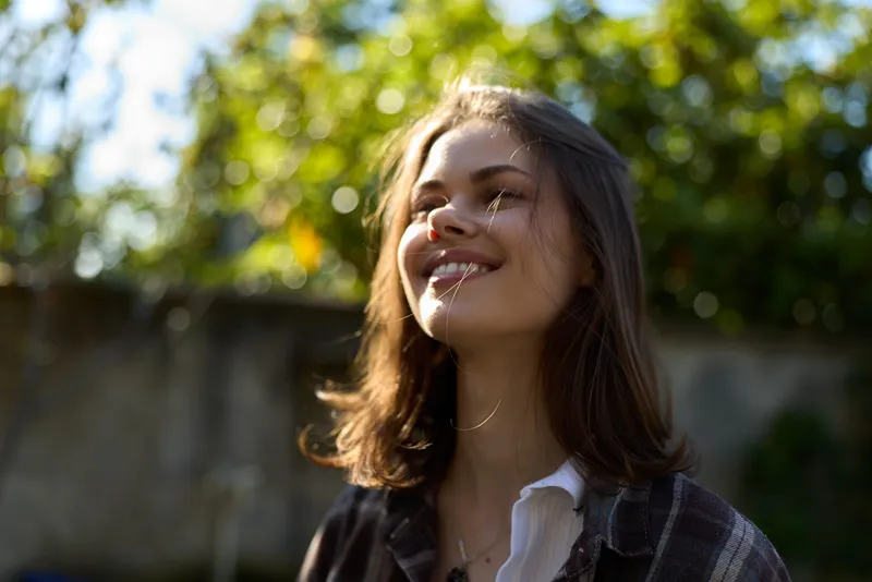 Bright energetic woman outdoors, smiling in sunlight, casual outfit, relaxed mood, candid portrait in a garden, lifestyle moment captured with warmth and confidence