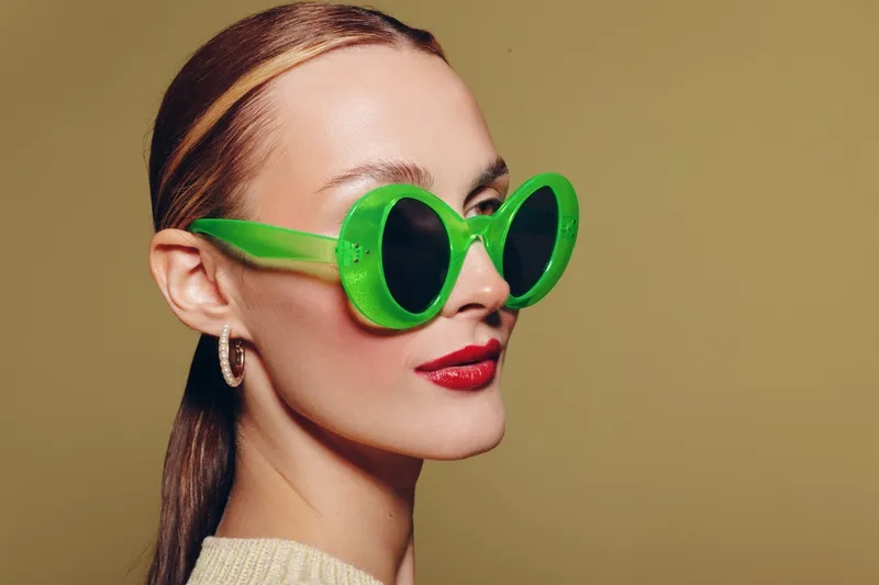 Stylish woman with slicked back hair wearing large green sunglasses and red lipstick poses against beige background. Concept of fashion, beauty, and modern accessories.