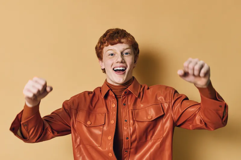 Young man with red hair wearing brown leather jacket smiling and raising fists celebrating success in front of beige background with positive energy and enthusiasm.