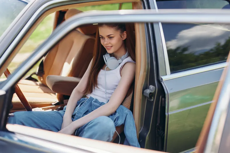 Young woman sitting in a vintage car, wearing casual denim and headphones, enjoying a sunny day with a relaxed and cheerful expression, outdoors.