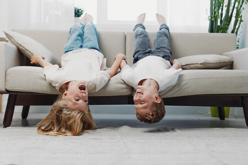 Two happy children hang upside down from sofa laughing and holding hands in bright living room. Concept of childhood joy, friendship, and playful moments.