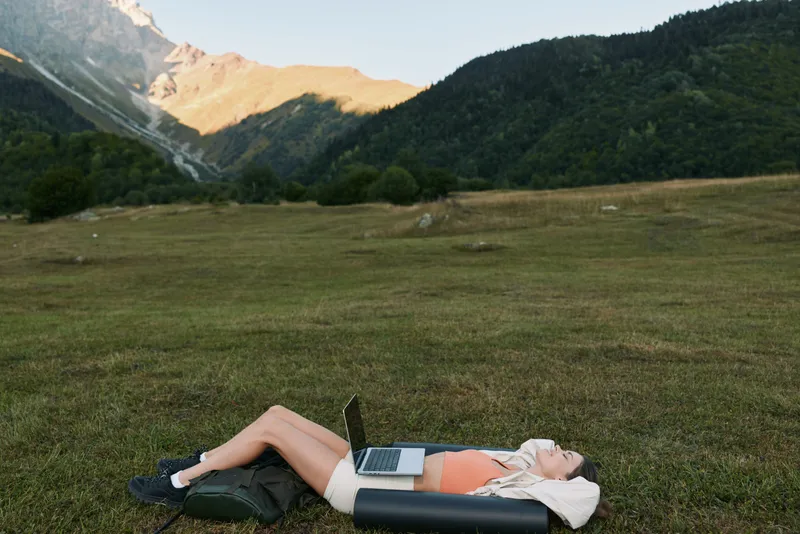 A relaxed entrepreneur lies on a grassy field with a laptop resting on her chest, mountains in the background, and warm sunlight creating a calm, productive outdoor mood.