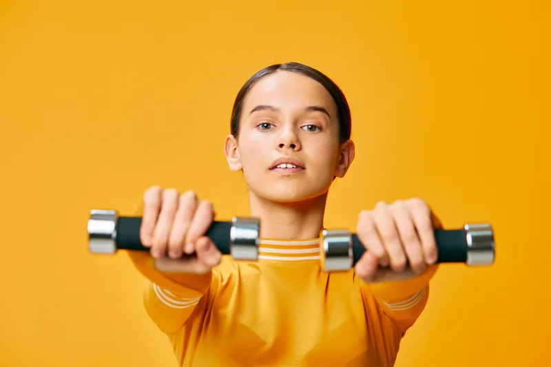 A focused woman fitness portrait lifting dumbbells in a studio sport scene, yellow top against vibrant orange background. Closeup of arms extended, strength and healthy lifestyle concept.