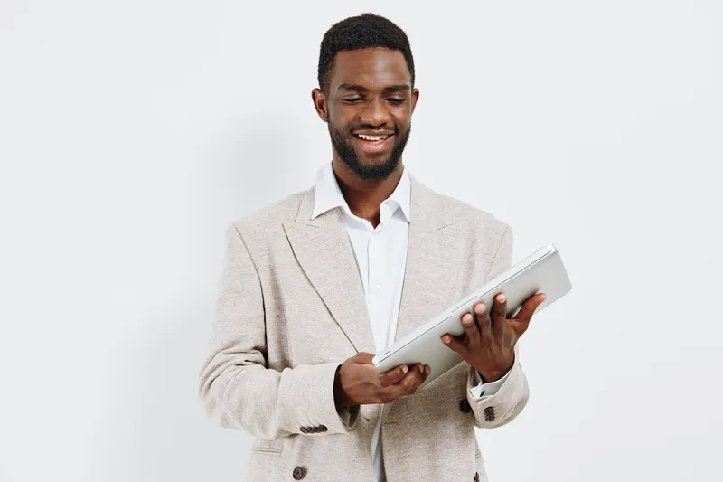 Smiling man wearing beige blazer and white shirt holding tablet device in hands, looking at screen isolated on plain white background, studio portrait with a happy expression, casual business look. Pe