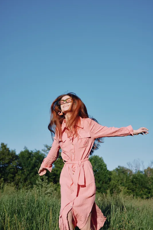 Joyful young woman in a pink dress dancing in a sunny field, showcasing carefree emotions, vibrant green background, and clear blue sky