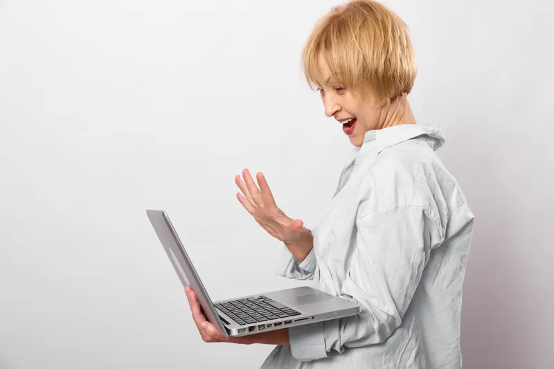 Woman with short blond hair holding a laptop, looking stressed and waving her hand near the keyboard. Indoor studio portrait on white background, technology and communication