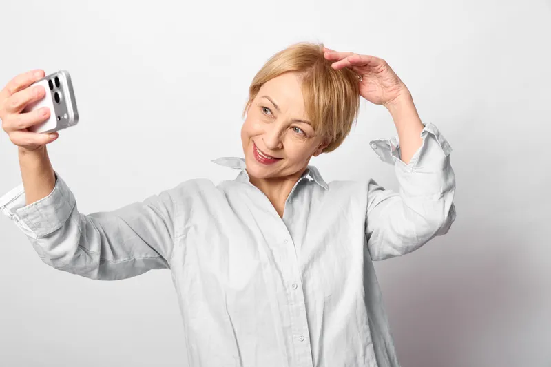 Stressed woman selfie with smartphone, anxiety in blonde portrait, hand on head with headache expression, older casual shirt, white indoor background, mental stress concept today